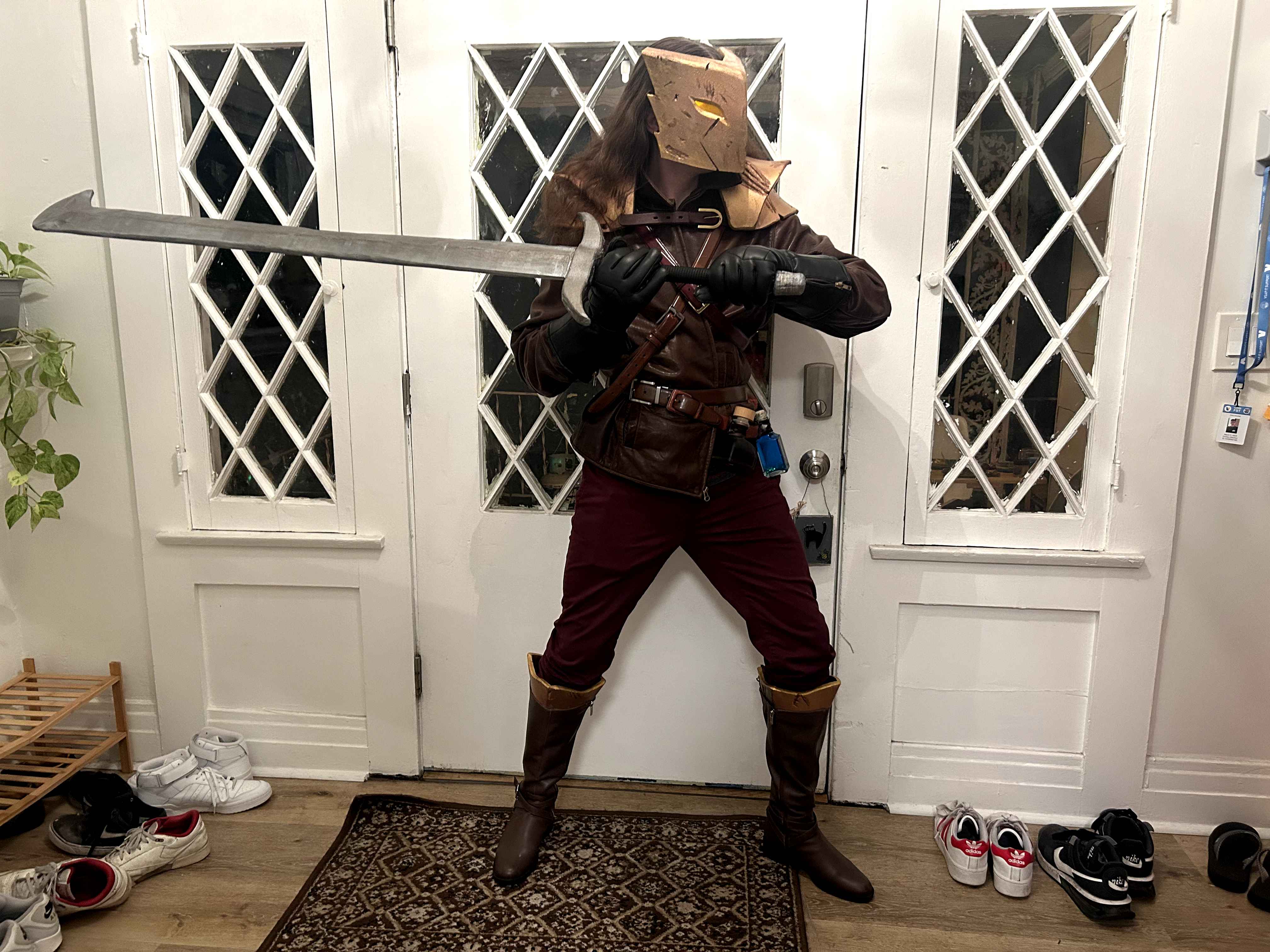 A cosplay of The Ironclad with a leathery vest, potion belt, shiny pauldrons and mask with glowing eyes, signature sword, red pants, and leathery boots, with the cosplayer in Ironclad’s iconic combat pose.